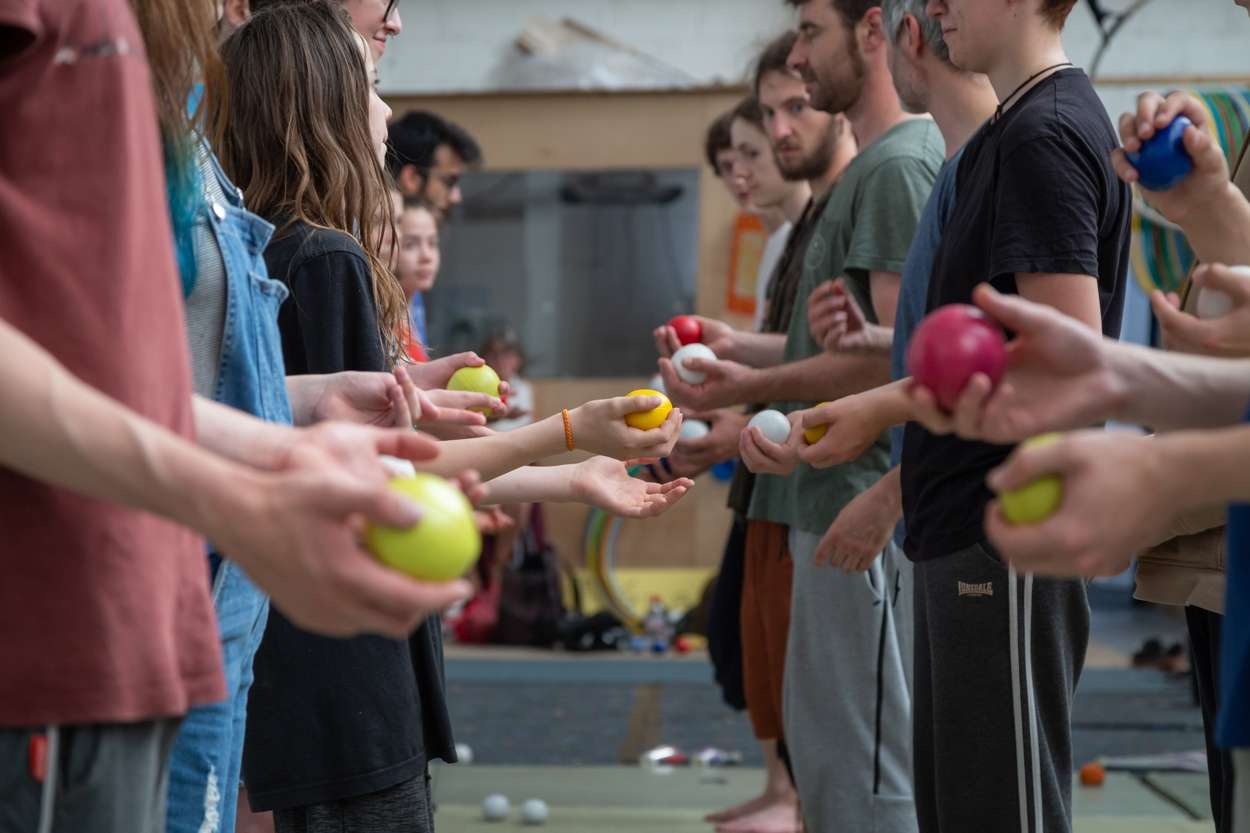 Deux rangées de personnes sont alignées en face à face avec les mains tendues et dans chacune d'entre elles, une balle colorée. 