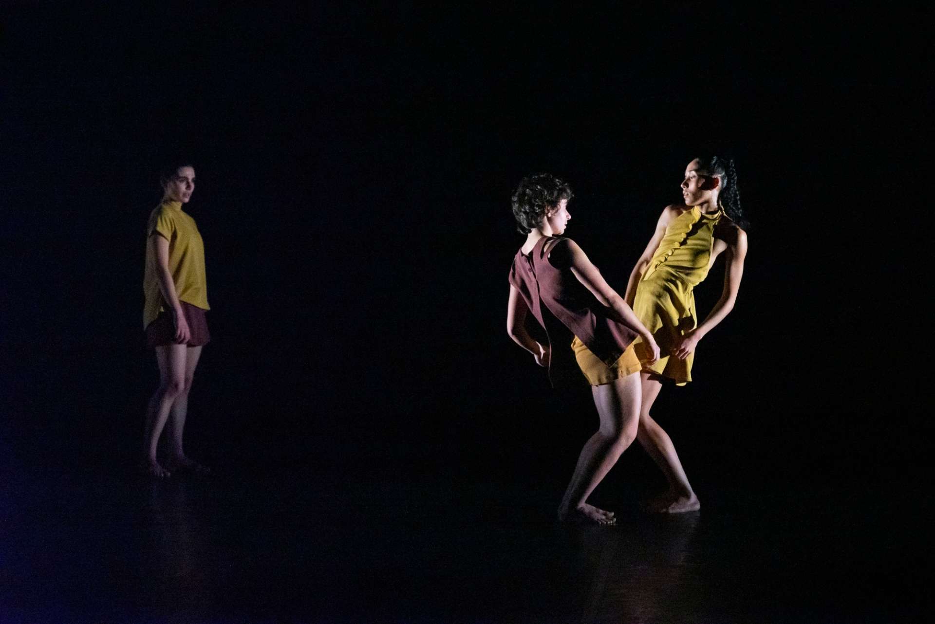 Sur fond noir, trois interprètes de l'ensemble chorégraphique du conservatoire de Paris dansent dans des tenues d'un jaune ocre. 