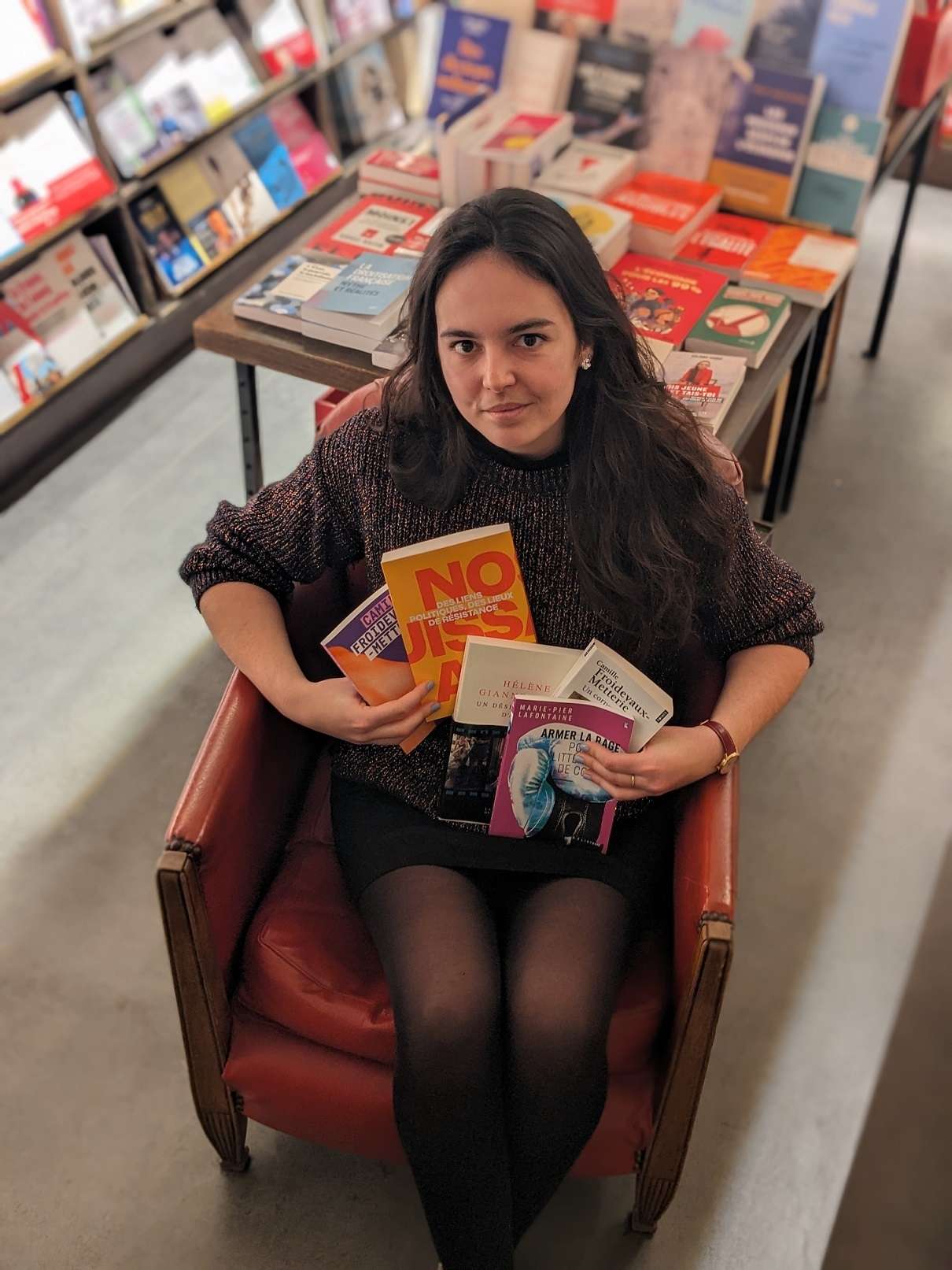 Deborah Vedel, libraire chez Les Temps Modernes à Orléans, est assise sur un fauteuil et tient des livres dans ses bras.