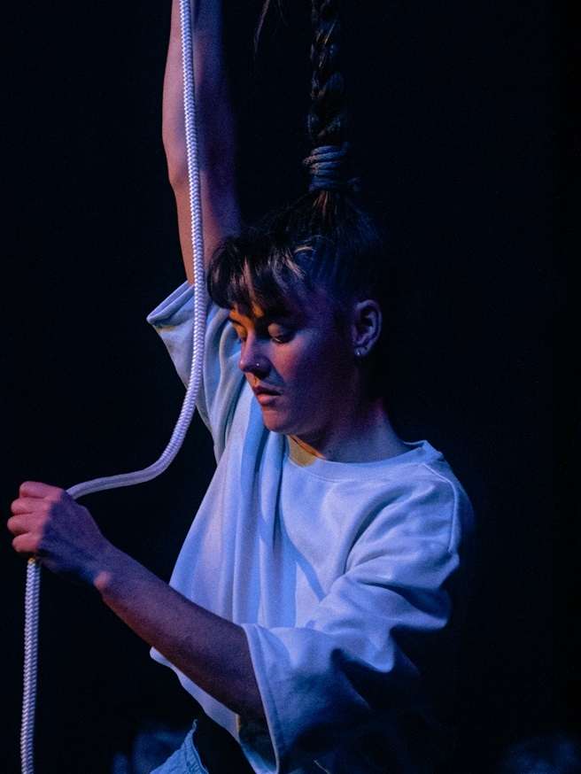 Une femme tient dans ses mains une corde suspendue, ses cheveux, coiffés en nattes, sont accrochés à cette corde. 